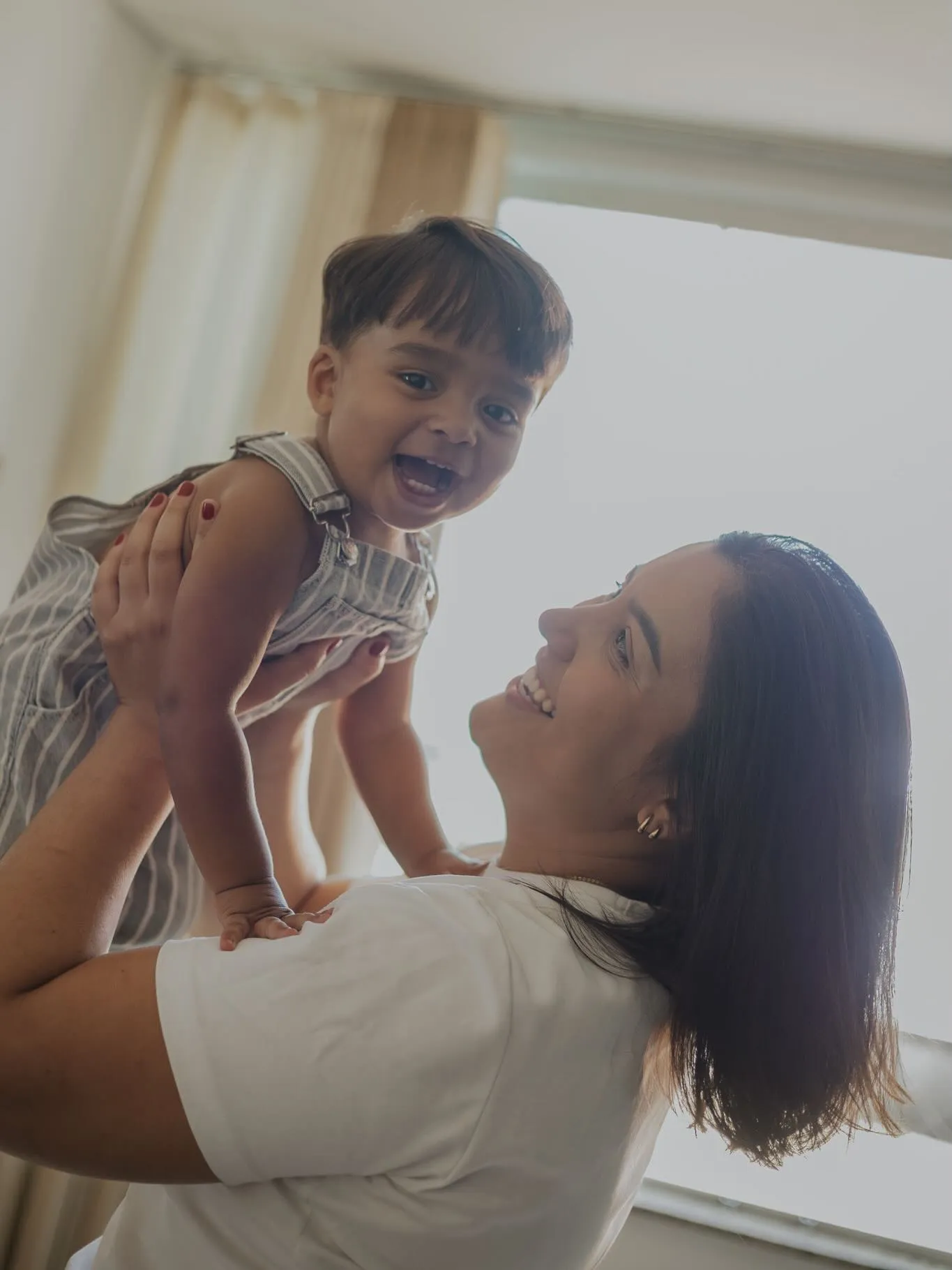 Mãe levanta o filho no colo, os dois rindo, em casa