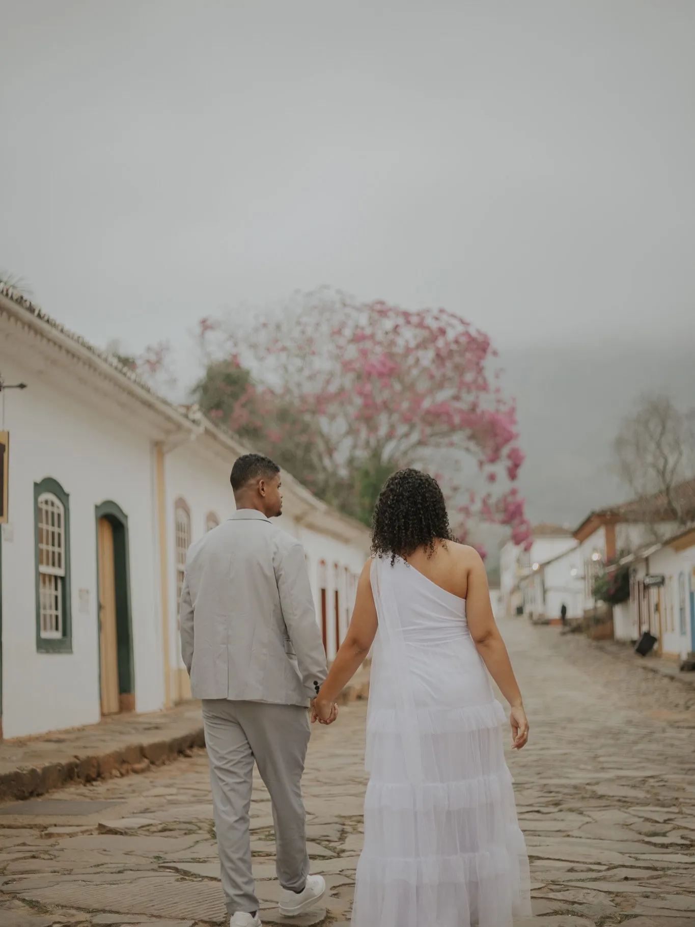 Casal de mãos dadas caminhando por rua de pedra em Tiradentes, com ipê florido ao fundo