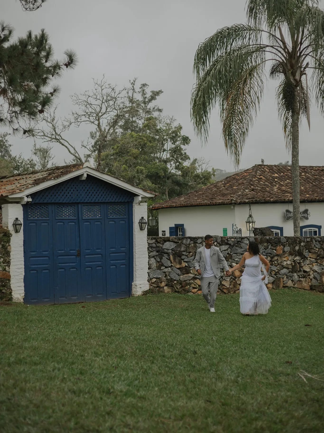 Casal correndo de mãos dadas em um gramado diante de casinha de porta azul